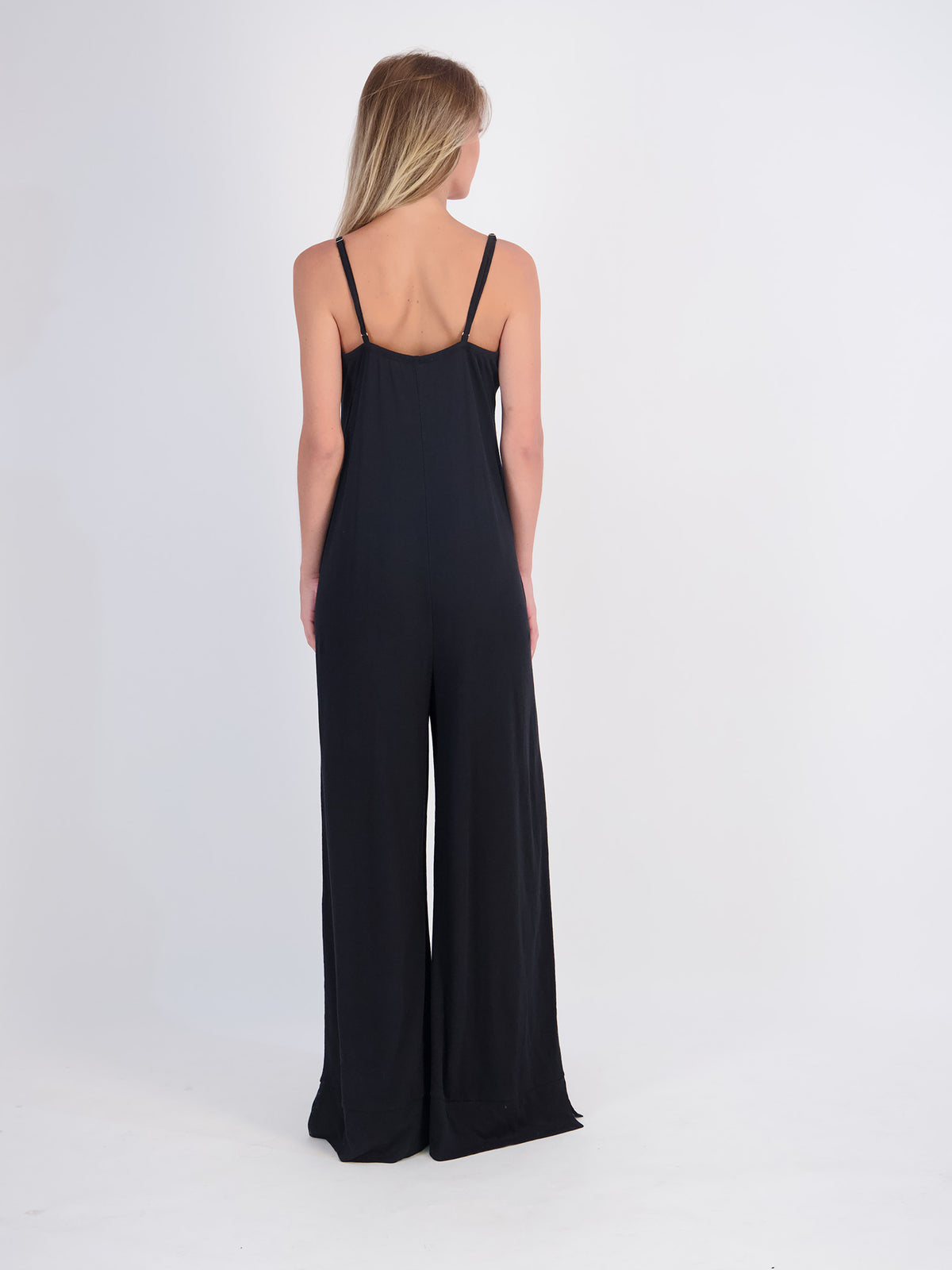 A woman facing backwards modeling a black sleeveless jumpsuit with wide legs, standing against a plain white studio background.